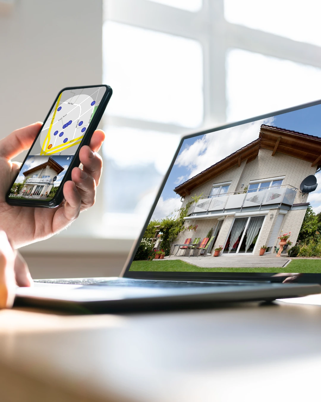 Person holds a phone showing a property map with blue pins while a laptop screen displays a two‑story house with a patio.