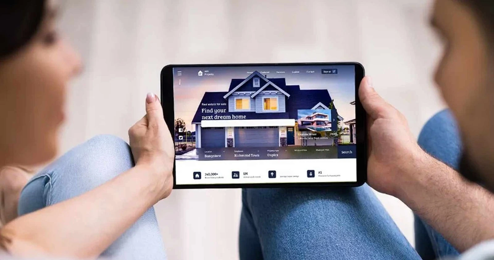Two people sit with a tablet displaying a real estate website featuring a large house image and search options nearby