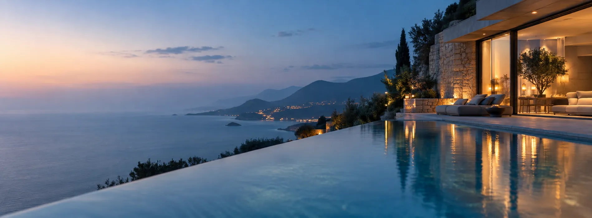 An infinity pool at a hillside villa at blue hour, lights glowing, the sea and coastline beyond