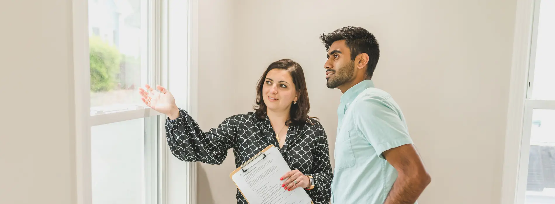 A real estate agent with a clipboard points out the window to her client in a bright, empty home