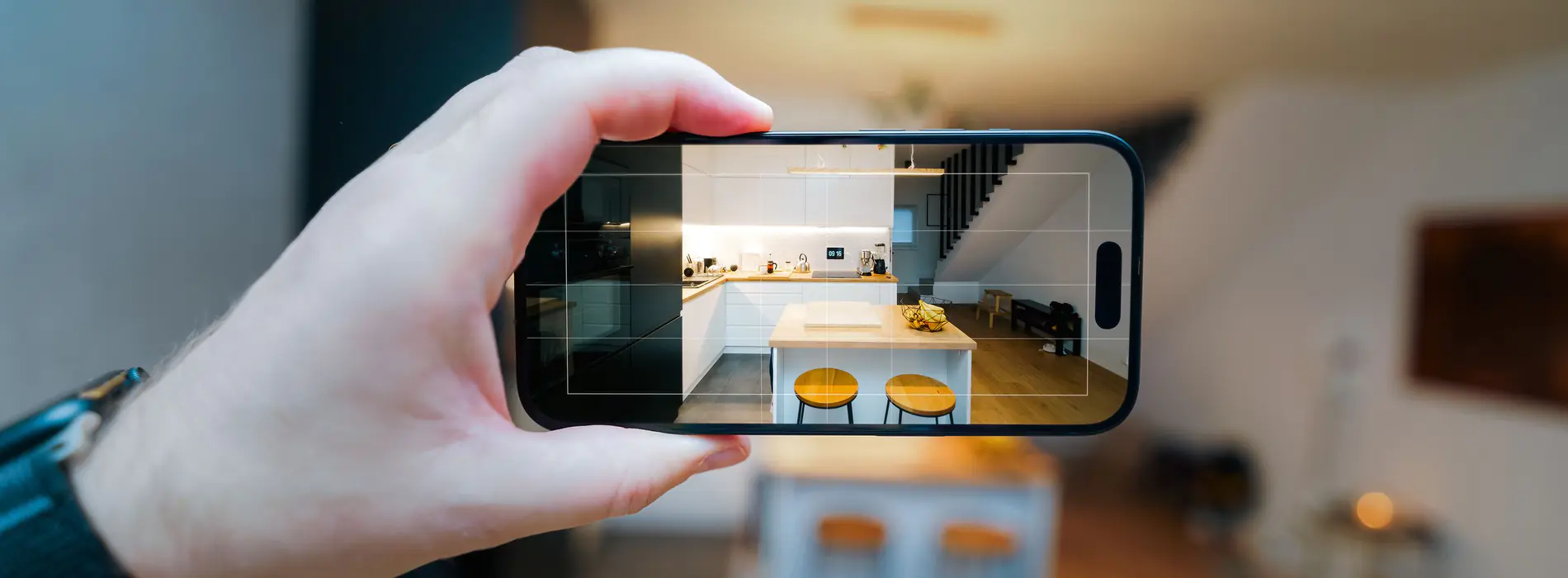 A hand holding a phone sideways to take a real estate listing photo of a modern white kitchen with a wooden-topped island and two yellow stools