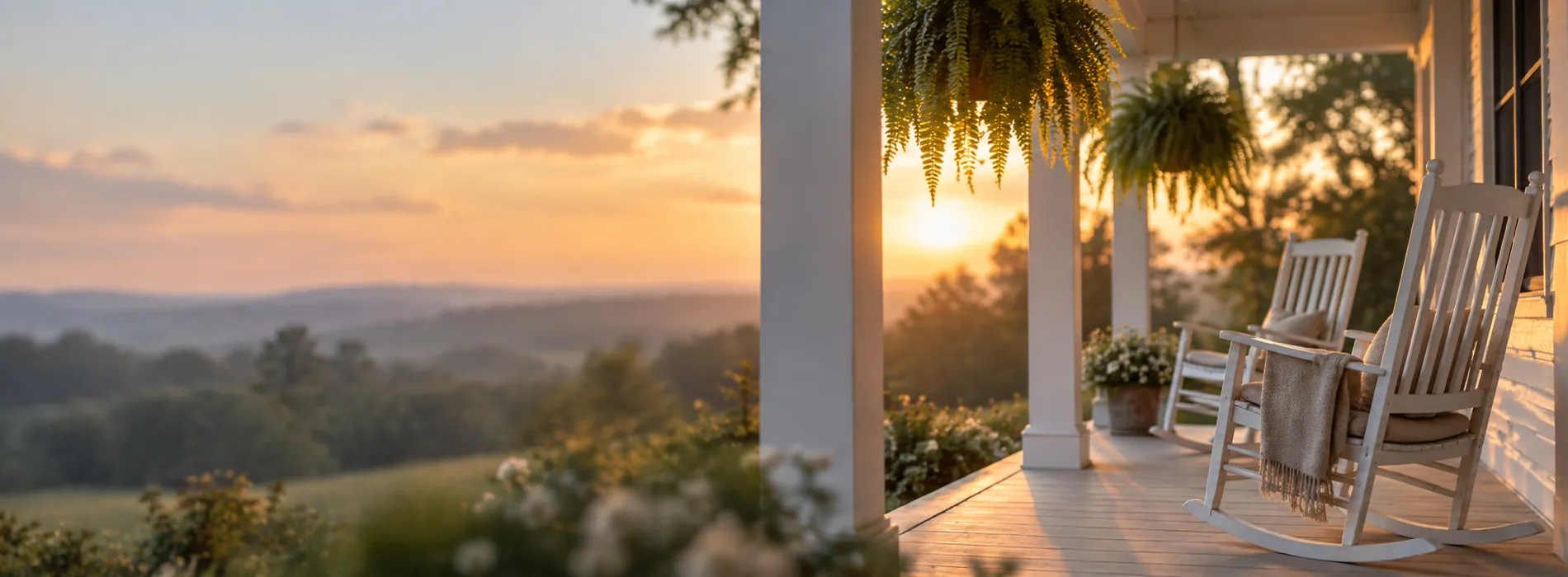 A white porch with rocking chairs and hanging ferns at sunset over green hills