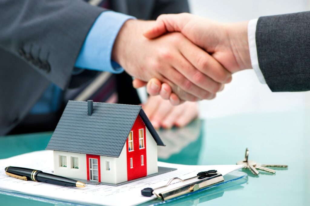 Handshake between two professionals over a miniature house model on a desk with documents, a pen, and keys nearby.
