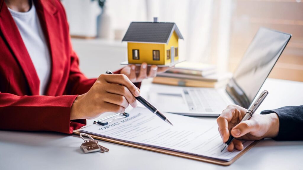 Two people sign a real estate contract at a desk, with a yellow house model and keys nearby on the table.