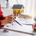 shutterstock_2285393485-scaled-1 - Kihan Marketing Two people sign a real estate contract at a desk, with a yellow house model and keys nearby on the table.