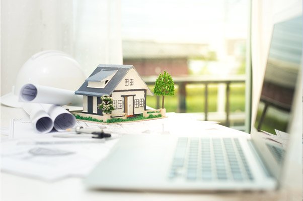 Small architectural model house on a desk with rolled blueprints, a laptop, a hard hat, and a tree, suggesting home design planning.