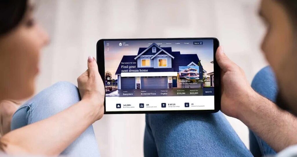 Two people sit with a tablet displaying a real estate website featuring a large house image and search options nearby