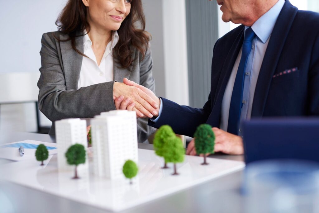 Two professionals shake hands over a desk with a scale model of buildings and trees, signaling a business agreement in real estate planning