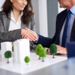 close-up-business-partners-shaking-hands-scaled-1 - Kihan Marketing Two professionals shake hands over a desk with a scale model of buildings and trees, signaling a business agreement in real estate planning