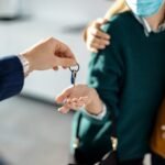 closeup-couple-receiving-key-their-new-real-estate-scaled-1 - Kihan Marketing A hand offers a key on a ring to another person, who is wearing a face mask.