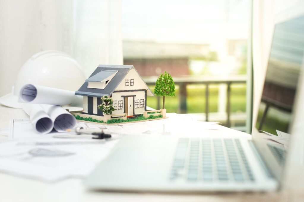 Miniature house model placed on architectural plans on a bright desk beside a laptop and rolled blueprints.