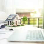 construction-concept-with-engineering-tools-scaled-1 - Kihan Marketing Miniature house model placed on architectural plans on a bright desk beside a laptop and rolled blueprints.