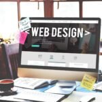 custom-real-estate-website-design - Kihan Marketing Desktop monitor showing a web design homepage with sticky notes, coffee cup, and sketches on a bright desk.
