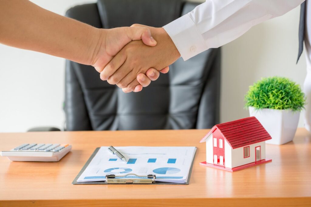 Two people shake hands over a desk with a chart, calculator, and a small red house model nearby.