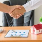 hands-agent-client-shaking-hands-after-signed-contract-buy-new-apartment-scaled-1 - Kihan Marketing Two people shake hands over a desk with a chart, calculator, and a small red house model nearby.