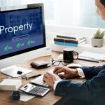house-loan-estate-sell-mortgage-concept-scaled-1 - Kihan Marketing Businessperson at a desk using a computer that shows a real estate portal with a large 'Property' header and green checkmarks for Buy, Sell, Rent