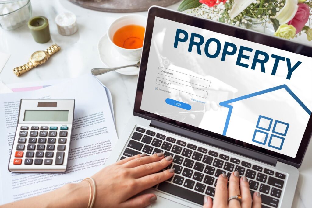 Laptop screen shows a property login form while a person types, with a calculator, papers, a cup of tea, and a vase of flowers on a white desk behind it.