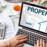 mortgage-house-loan-website-login-graphic-concept-scaled-1 - Kihan Marketing Laptop screen shows a property login form while a person types, with a calculator, papers, a cup of tea, and a vase of flowers on a white desk behind it.