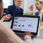 multi-ethnic-business-team-sitting-table-office-center-speaking-about-project-meeting-broadroom-scaled-1 - Kihan Marketing Business colleagues discuss data in a meeting, with a laptop showing charts and graphs on the screen.