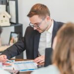 pexels-rdne-8293744-scaled-1 - Kihan Marketing Man in a suit and glasses explains a chart to two coworkers at a meeting table in an office.