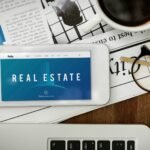 real-estate-accommodation-property-investment-graphic-word-scaled-1 - Kihan Marketing Tablet shows a real estate website on a blue banner, beside a cup of coffee and glasses on a newspaper.