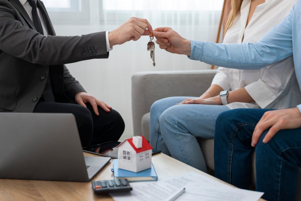 Two people pass keys to each other in a home-visit setting, with a model house and documents on the table nearby.