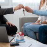 real-estate-agent-work-showing-house-doing-business-scaled-1 - Kihan Marketing Two people pass keys to each other in a home-visit setting, with a model house and documents on the table nearby.