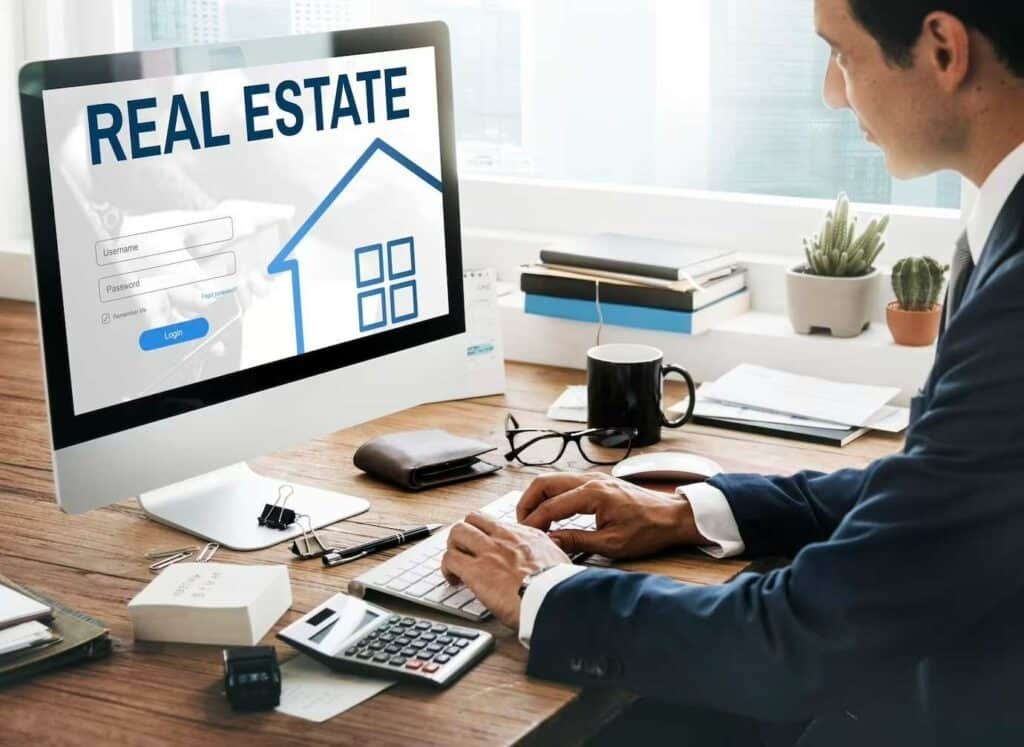 Man in a suit typing at a desk while a real estate website loads on a desktop monitor on his left side.