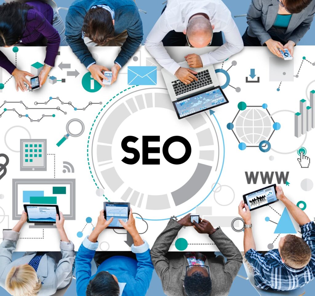 Top-down view of a diverse team gathered around a large SEO graphic, using laptops and tablets nearby.
