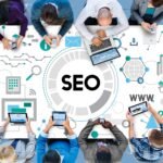 searching-engine-optimizing-seo-browsing-concept-scaled-1 - Kihan Marketing Top-down view of a diverse team gathered around a large SEO graphic, using laptops and tablets nearby.