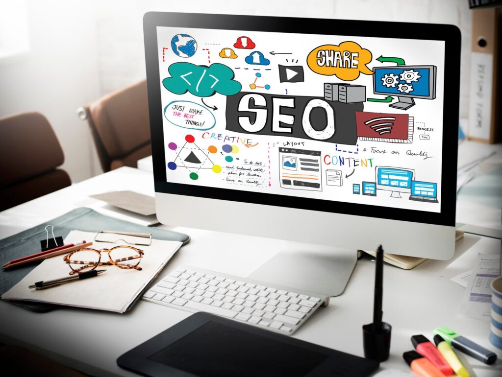 Monitor showing a colorful SEO doodle illustration on a white desk, with the word 'SEO' and related icons surrounding it.