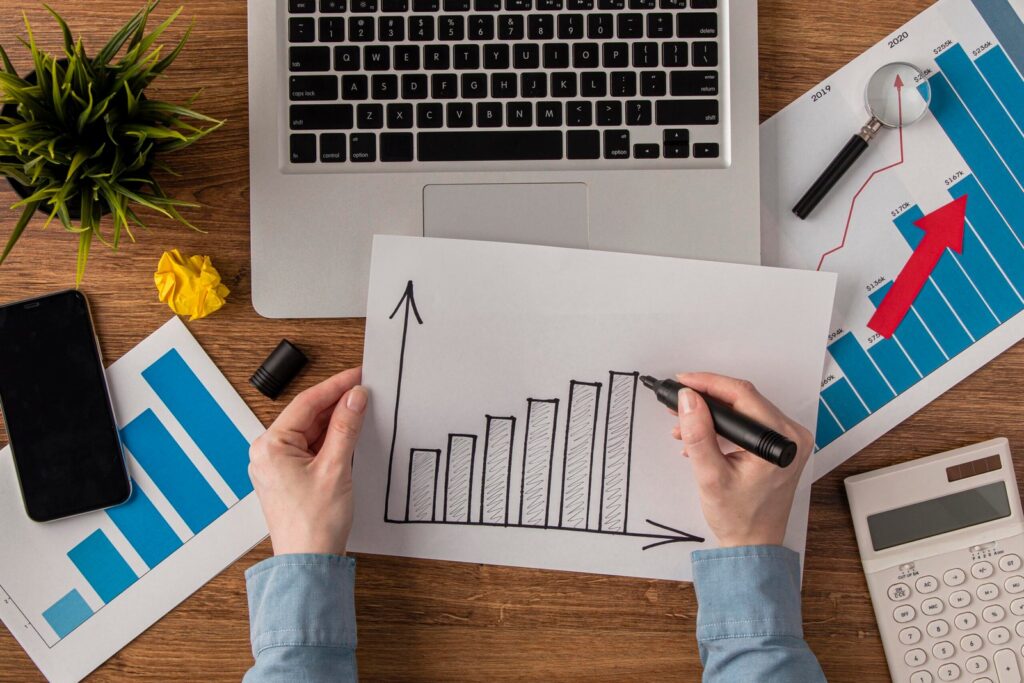 Hands draw an ascending bar chart on a white sheet at a wooden desk, with a laptop, papers, and a smartphone nearby.