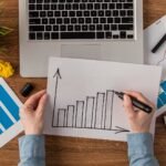 top-view-office-desk-with-calculator-growth-chart-scaled-1 - Kihan Marketing Hands draw an ascending bar chart on a white sheet at a wooden desk, with a laptop, papers, and a smartphone nearby.
