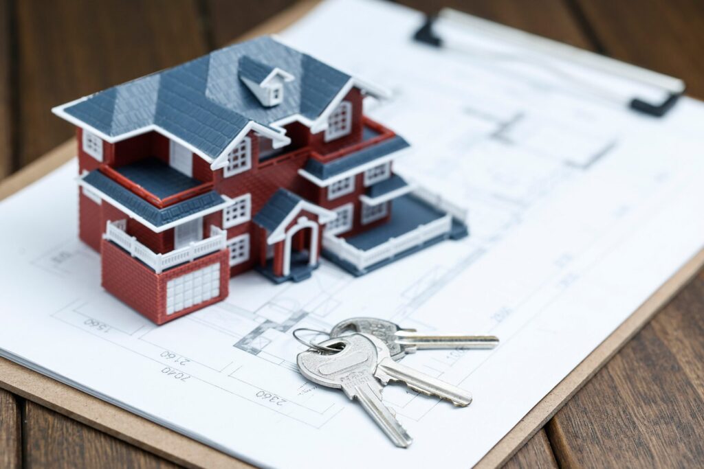 Model red-brick house figurine on architectural blueprints with a set of keys beside it, symbolizing real estate or home purchase