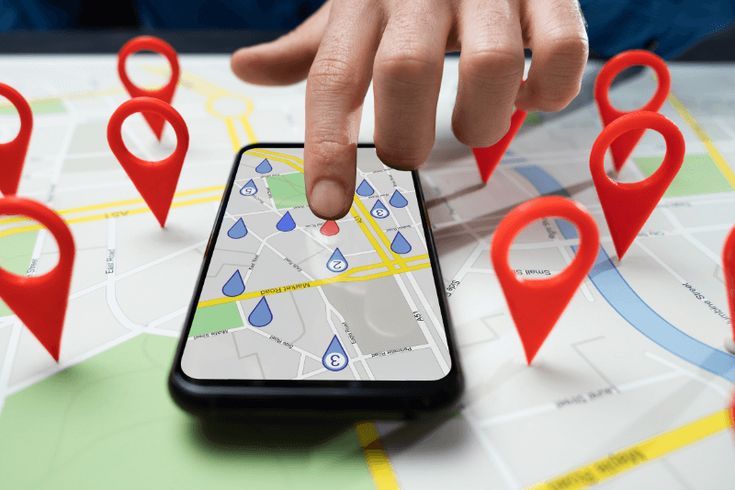 Hand taps a smartphone displaying a city map with multiple red location pins around it, indicating nearby points of interest.
