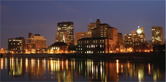 City skyline at dusk over a calm river, with illuminated windows and reflections on the water.