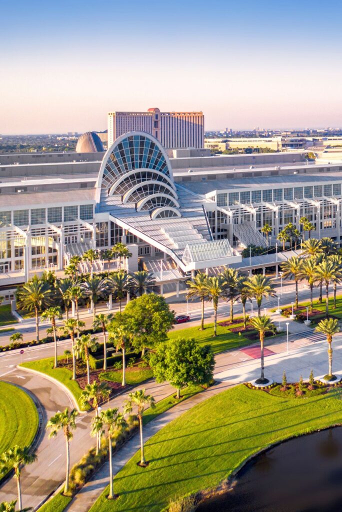 Modern convention center with curved glass arches, palm trees, and a waterfront lawn under a blue sky