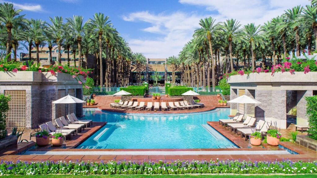 Resort pool with lounge chairs and white umbrellas, palm trees, and flowering planters along the deck.