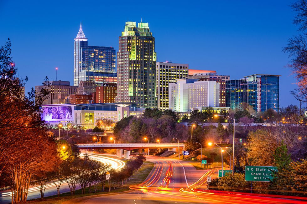 Evening city skyline with tall illuminated buildings and a busy highway with light trails in the foreground.