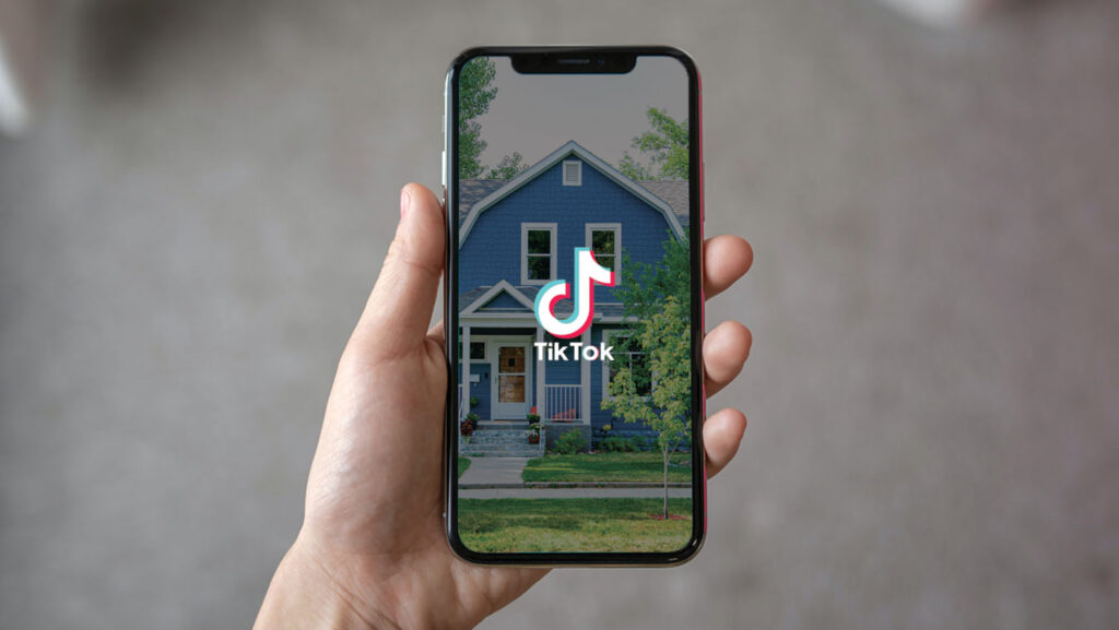 Hand holding a smartphone with the TikTok logo on the screen, over a blue house image in the background