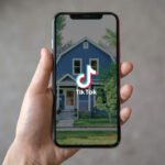 627d0b1c928ccc6f0cd3dffb_hero_image - Kihan Marketing Hand holding a smartphone with the TikTok logo on the screen, over a blue house image in the background