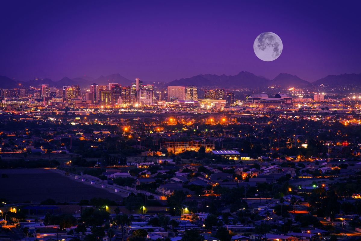 Nighttime city skyline with illuminated buildings and a full moon rising over distant mountains in a purple sky.