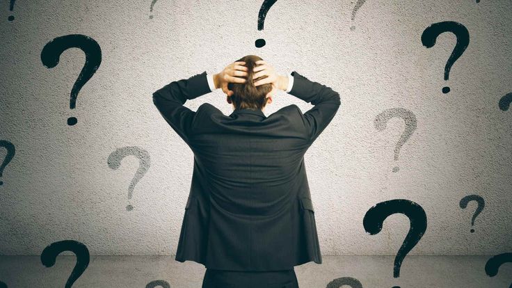 Man in a suit stands with hands on head, surrounded by large question marks, signaling confusion or stress in a work setting.