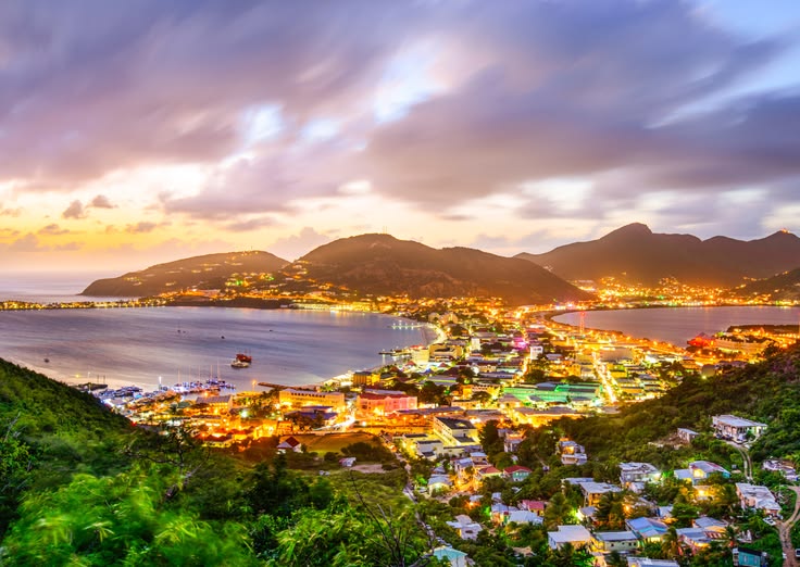 Coastal town glowing with colorful lights at dusk along a curved bay, hills in the background.