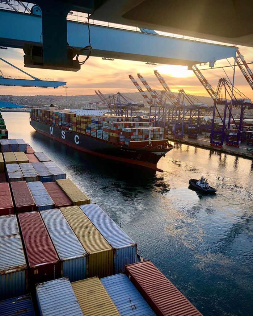 Container ship at a busy port during sunset, stacked with colorful cargo containers and large cranes overhead