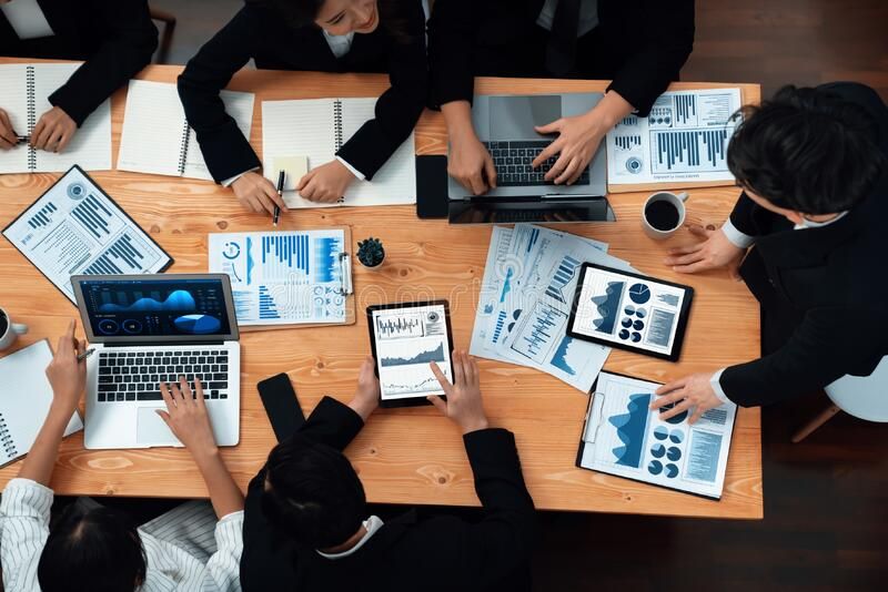 Top-down view of a team around a wooden table analyzing charts on laptops, tablets, and printed reports. Used for a business meeting or data review scenario.