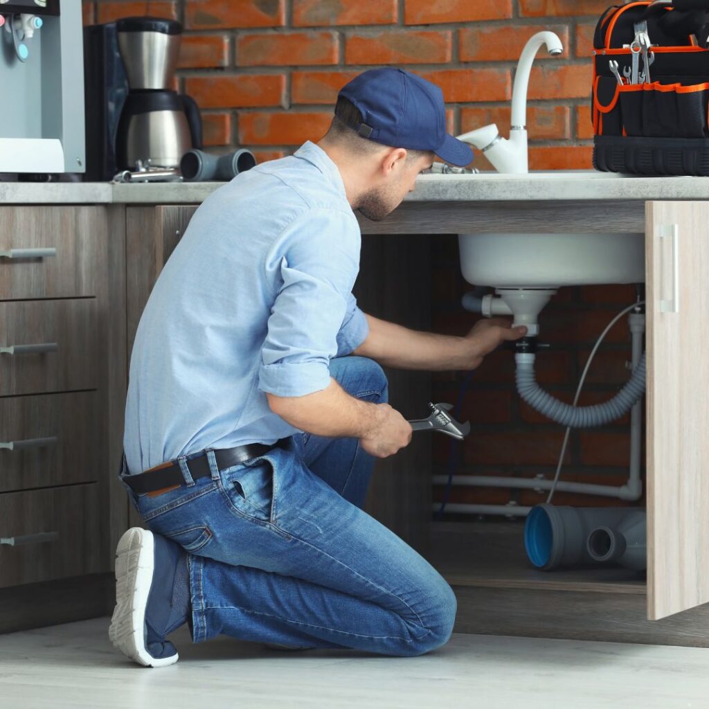 Plumber kneeling under a kitchen sink, tightening a pipe with a wrench under a cabinet.