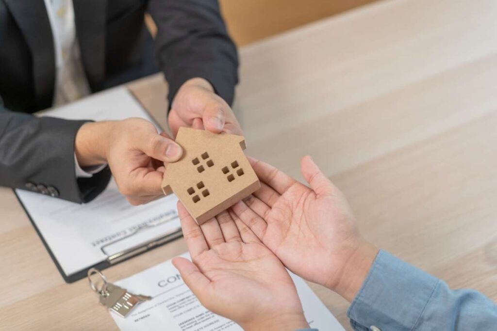 Two people exchange a small wooden house model over a table with contract documents nearby, symbolizing a real estate agreement at signing time.