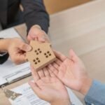 Asian young man client tenant buy home apartment - Kihan Marketing Two people exchange a small wooden house model over a table with contract documents nearby, symbolizing a real estate agreement at signing time.