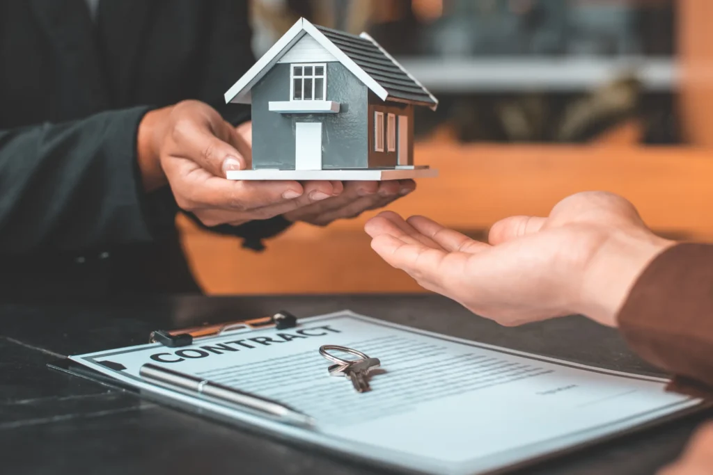 Hands exchanging a small house model over a signed contract on a table, with a key nearby (real estate transaction).